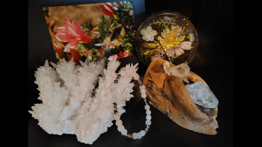 Resin memorial coaster with gold dragonflies, flowers, stones, and soil from Forever And A Day alongside aragonite crystal, photograph, bracelet and shamanic tool on black background